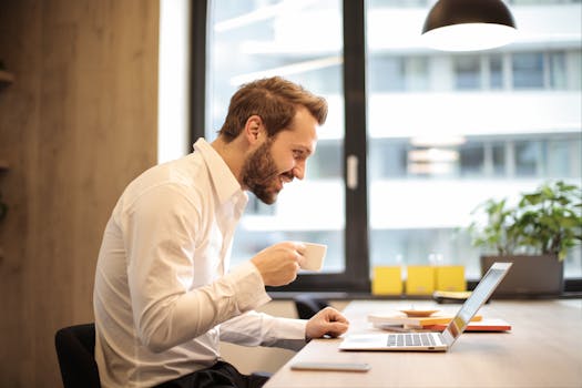 pexels-photo-925786-925786-2 Man in office with coffee, smiling while working at laptop, captures the essence of remote work.